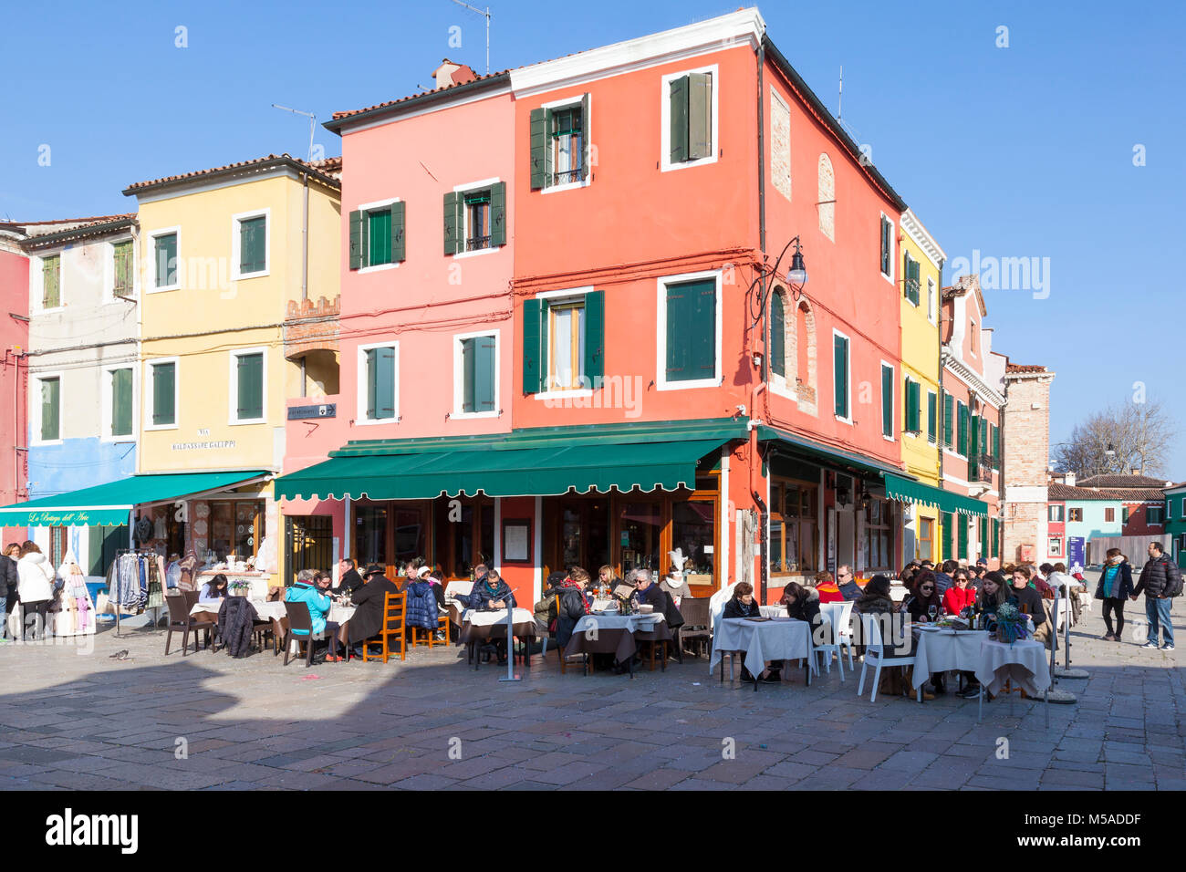 Burano Island, Venice, Veneto, Italy. People dining at an open-air restaurant in Via Baldassare Galuppi in winter with bright colourful buildings Stock Photo