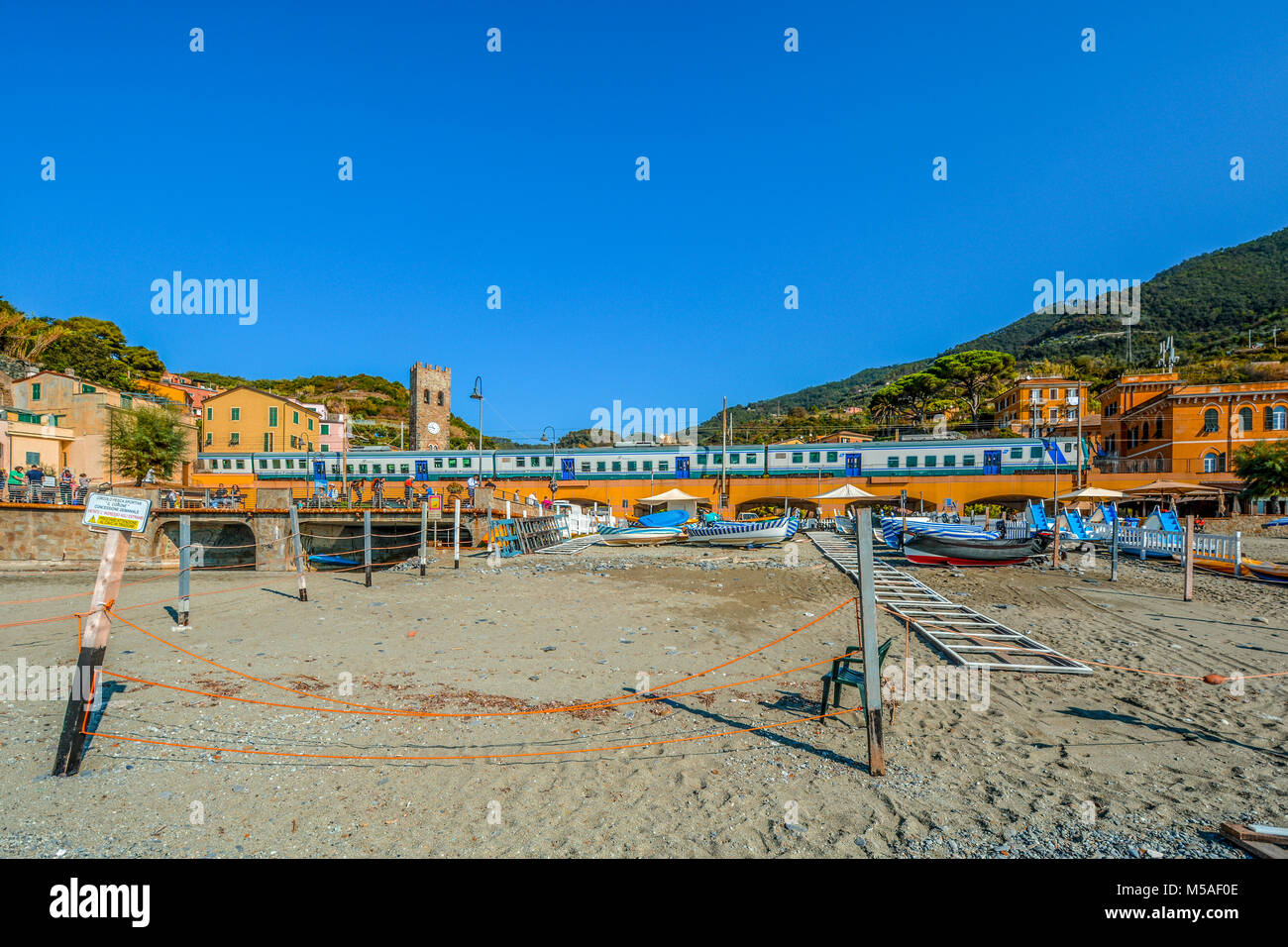 The Trenitalia train travels through the Cinque Terre village of Monterosso Al Mare with the town including church clock tower and the sandy beach Stock Photo