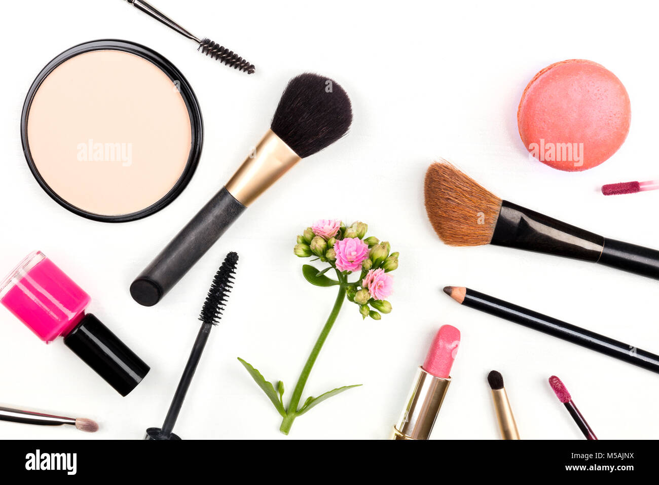 A closeup of makeup brushes, powder, lipstick, and a mascara applicator, shot from above on a white background with a pink flower and a macaroon, with Stock Photo