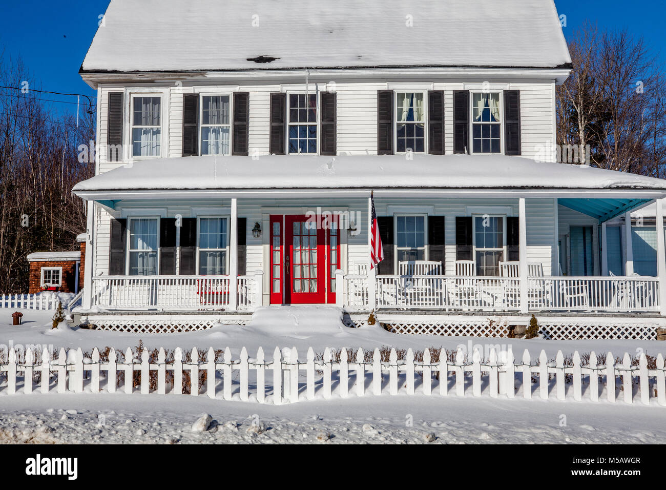 A traditional white-painted New Englander home with a wrap-around porch, red front door, black shutters, American Flag, picket fence and a yard full o Stock Photo