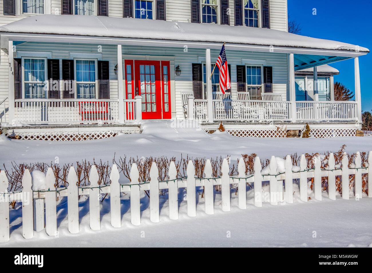 Facade of a traditional, white-painted New Englander home with an American Flag, red door, picket fence and wrap-around porch with yard covered in dee Stock Photo