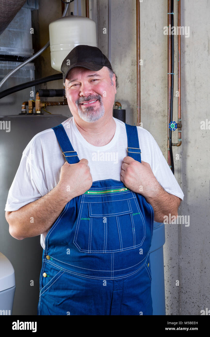 Trustworthy middle-class workman in a basement utility room standing holding his denim overalls as he smiles at the camera Stock Photo
