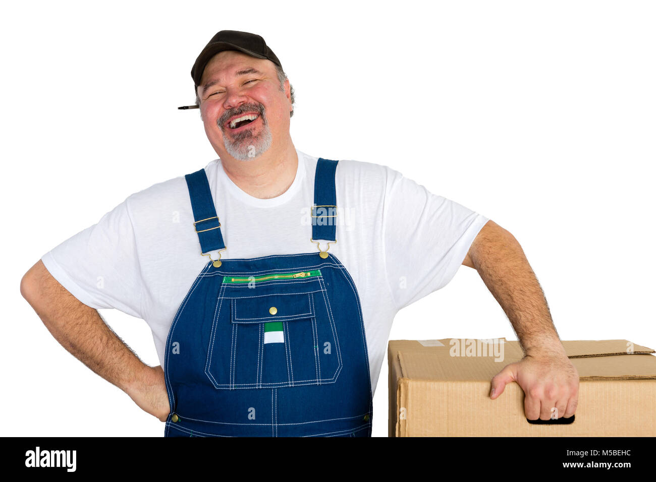 Cheerful delivery wearing dungarees man standing by big box against white background Stock Photo