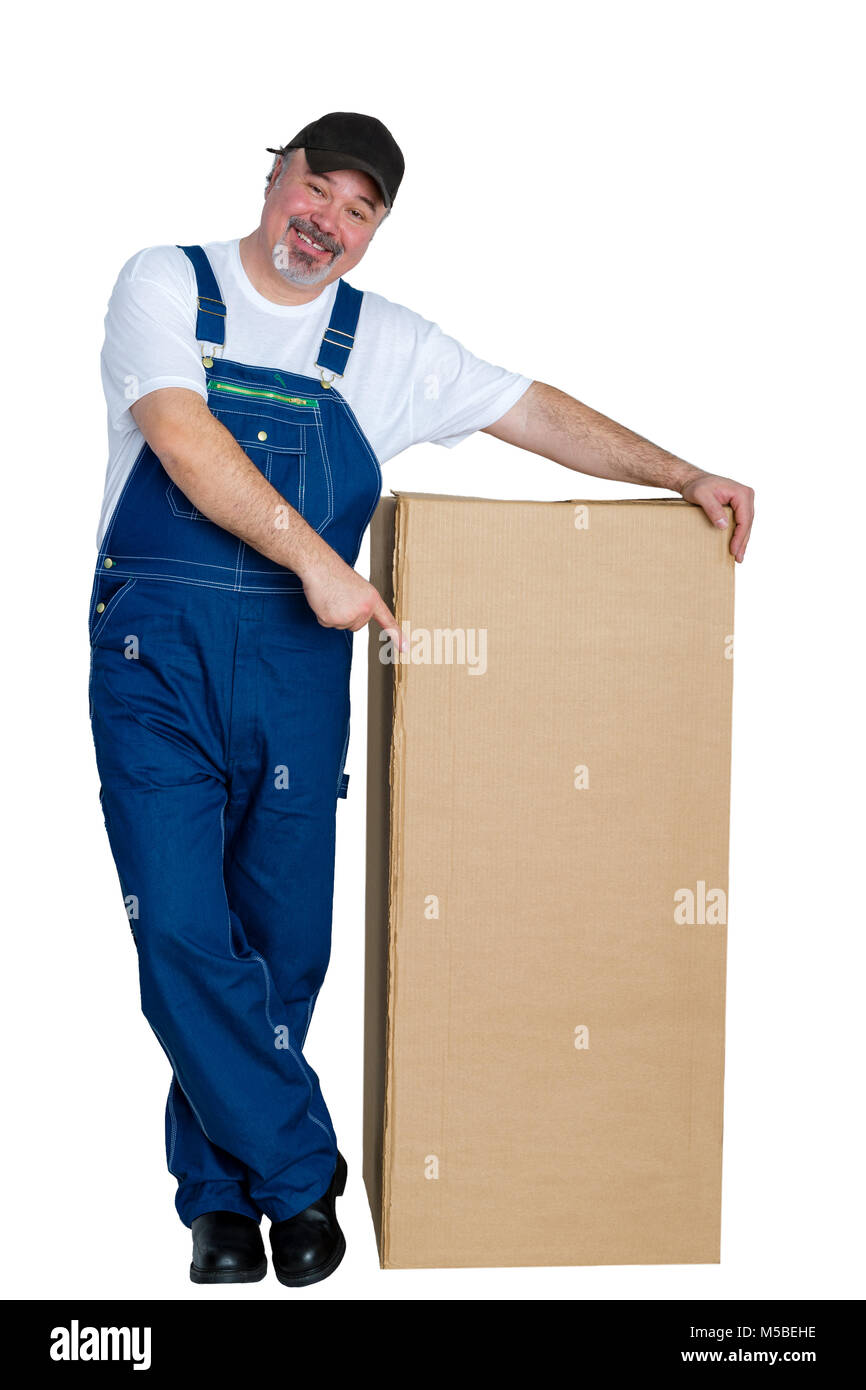 Smiling happy worker in denim overalls standing pointing to a large brown unlabelled cardboard box with copy space isolated on white Stock Photo