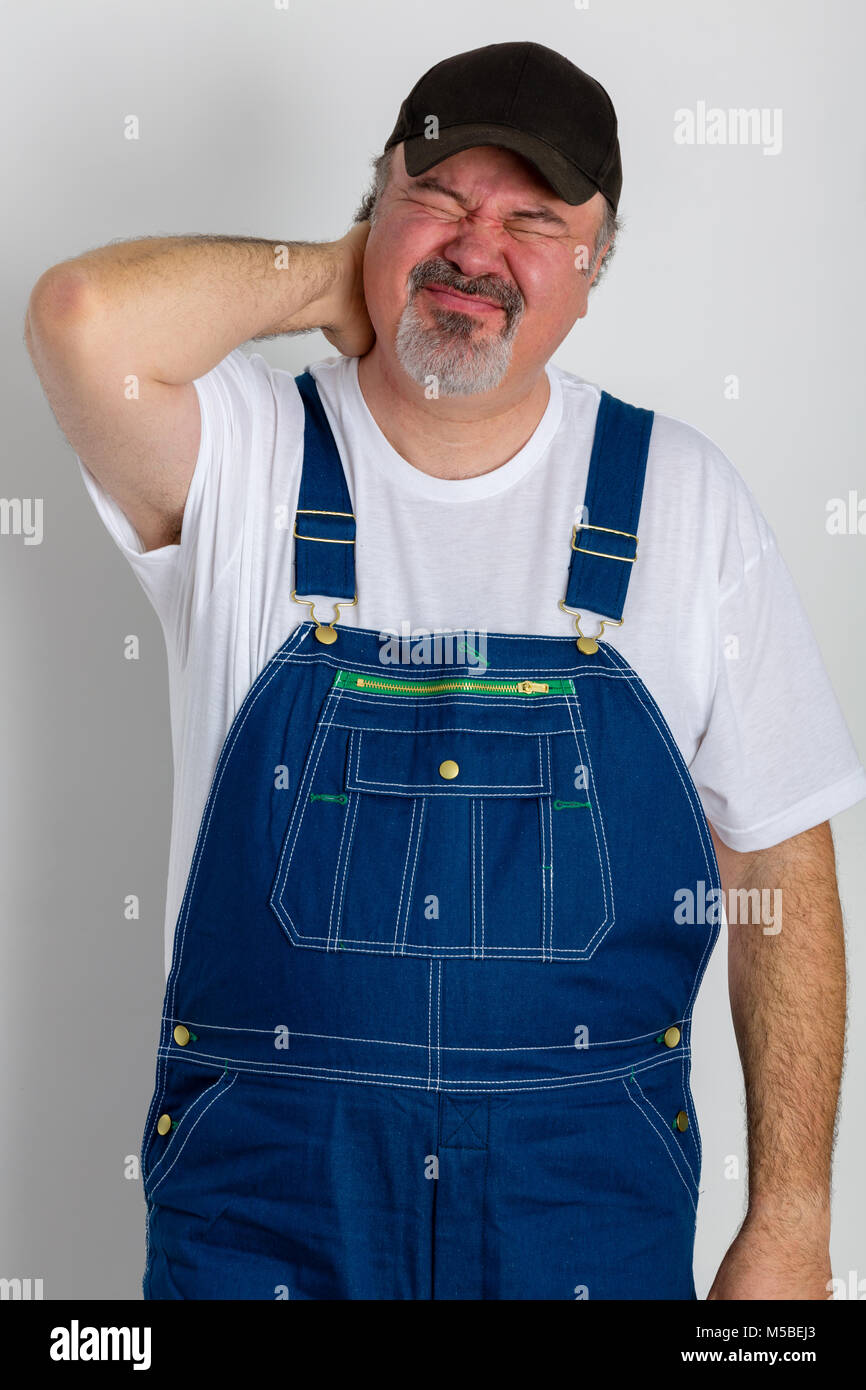 Mature man with grimace on face wearing dungarees holding his painful neck Stock Photo