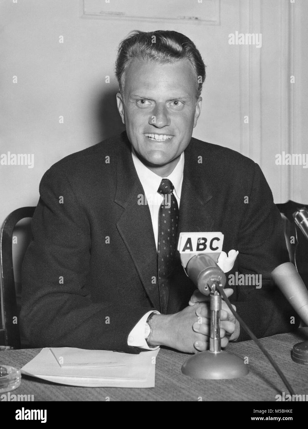 American evangelist Billy Graham (1918-2018) sitting at desk with media microphones on September 20, 1958. Stock Photo