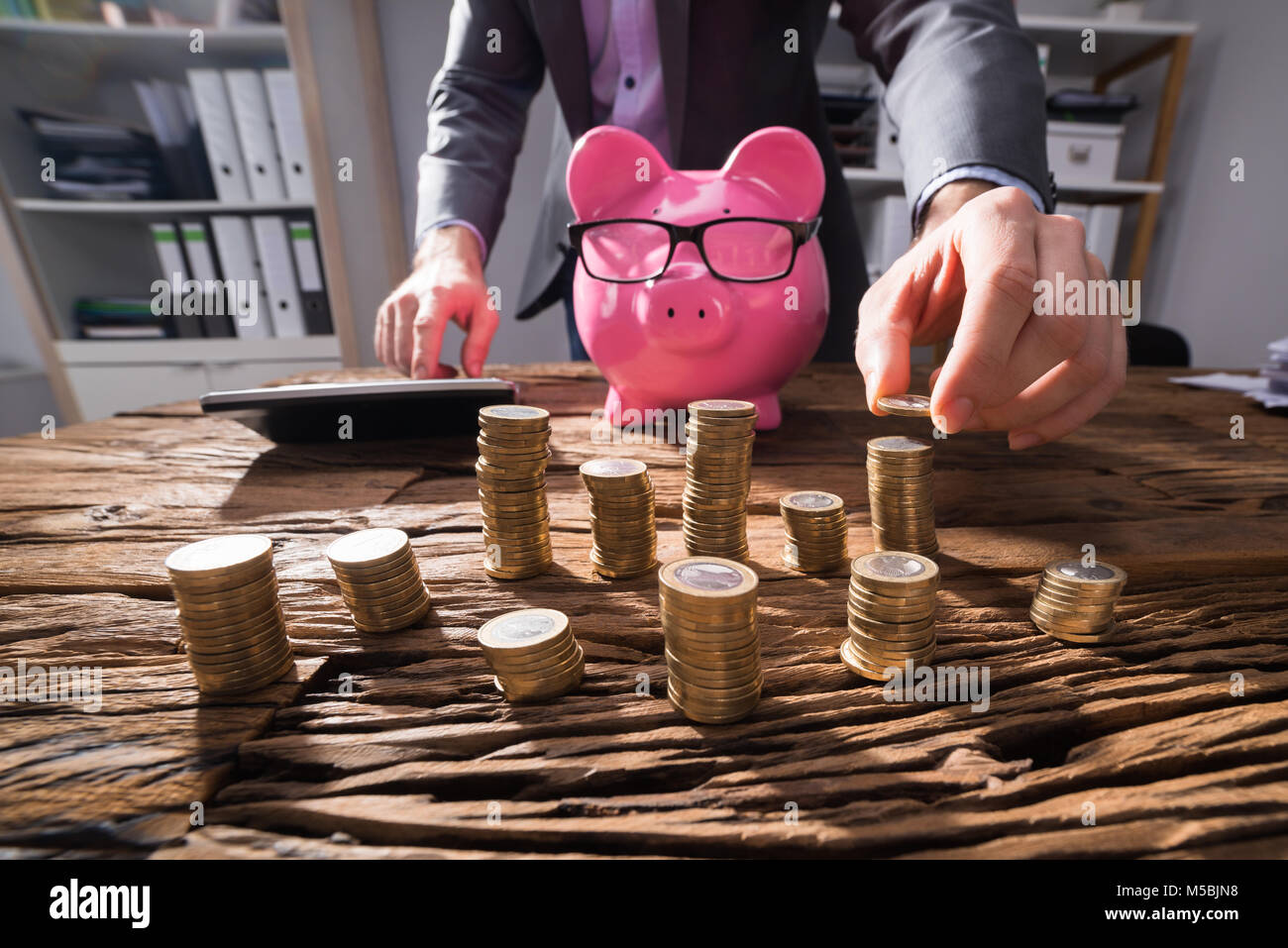 Businessperson Calculating Stacked Golden Coins With Pink Piggybank On Wooden Desk Stock Photo