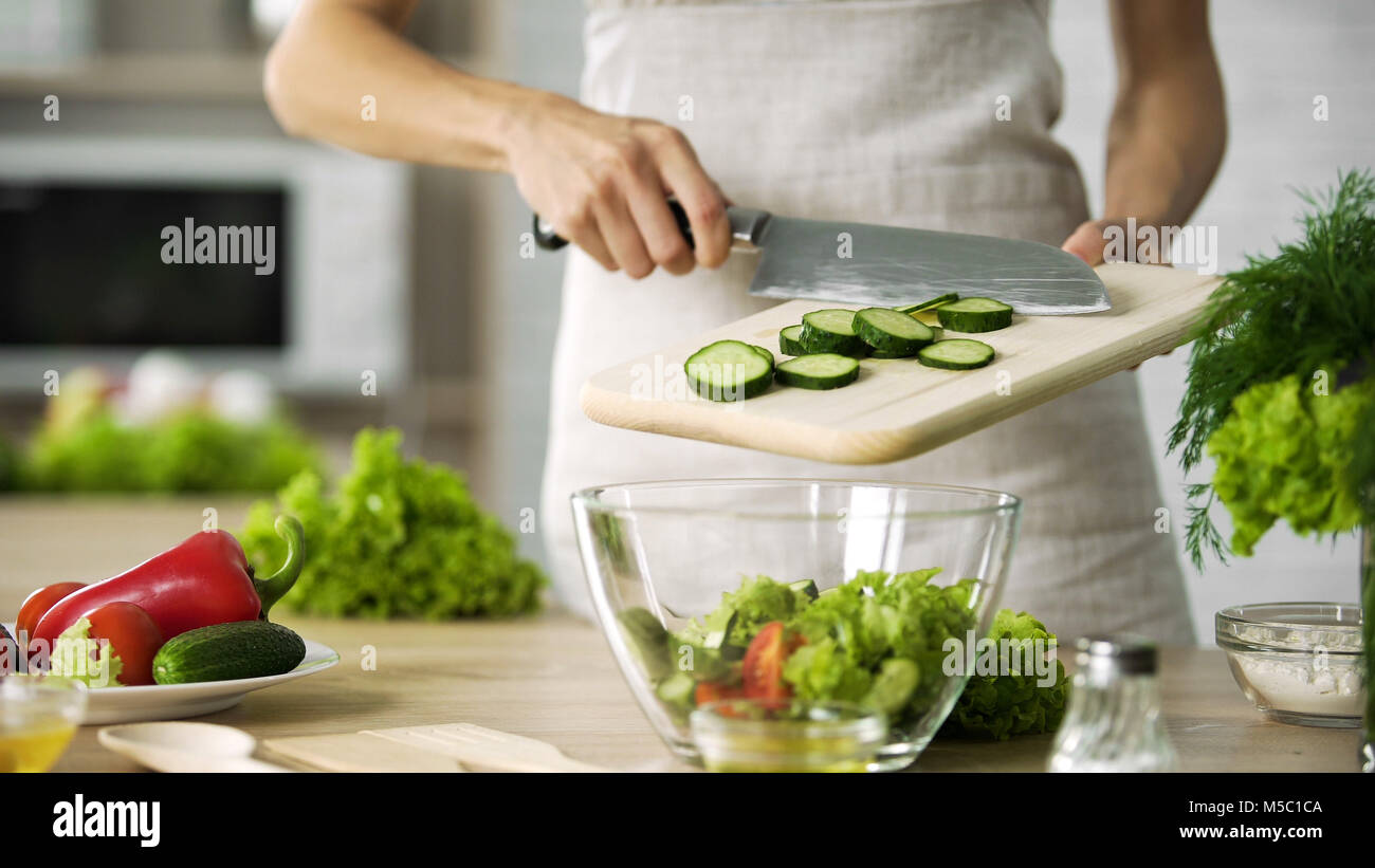 Professional female cook adding fresh cucumber slices in glass bowl ...