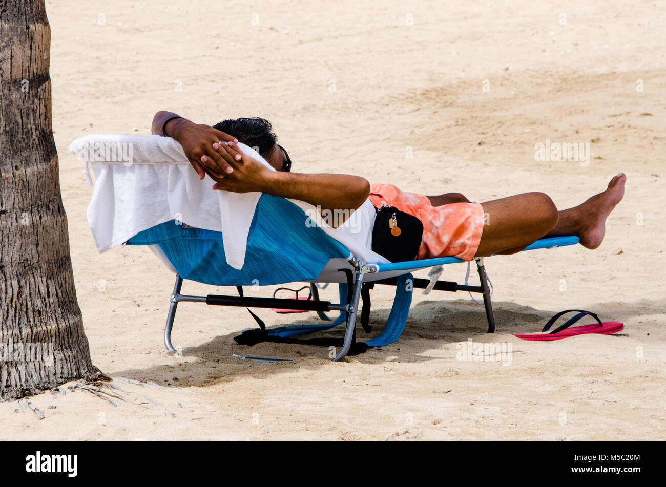 Male reclining on beach chair sunbathing under palm tree on beach. Man enjoying retirement and beach lifestyle relaxes while sit on deck chair. Stock Photo