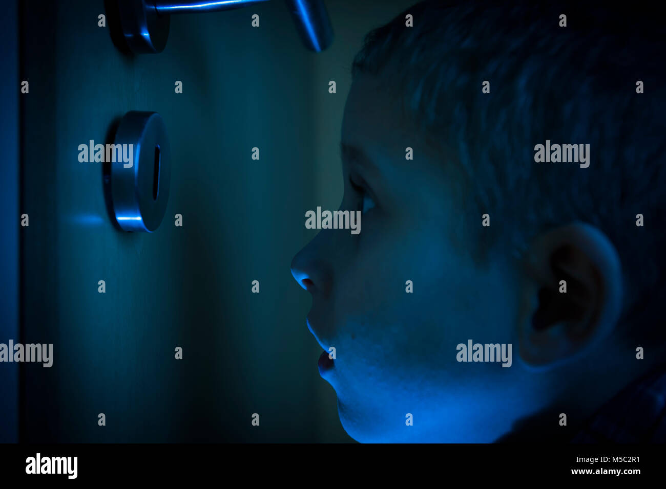 Junge im dunklen Zimmer blickt im Schein der Taschenlampe durch ein Schlüsselloch. Stock Photo