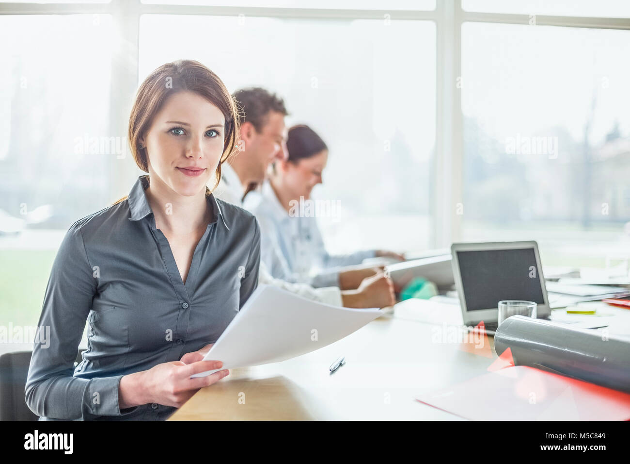 Portrait of beautiful young woman holding documents with colleagues in ...