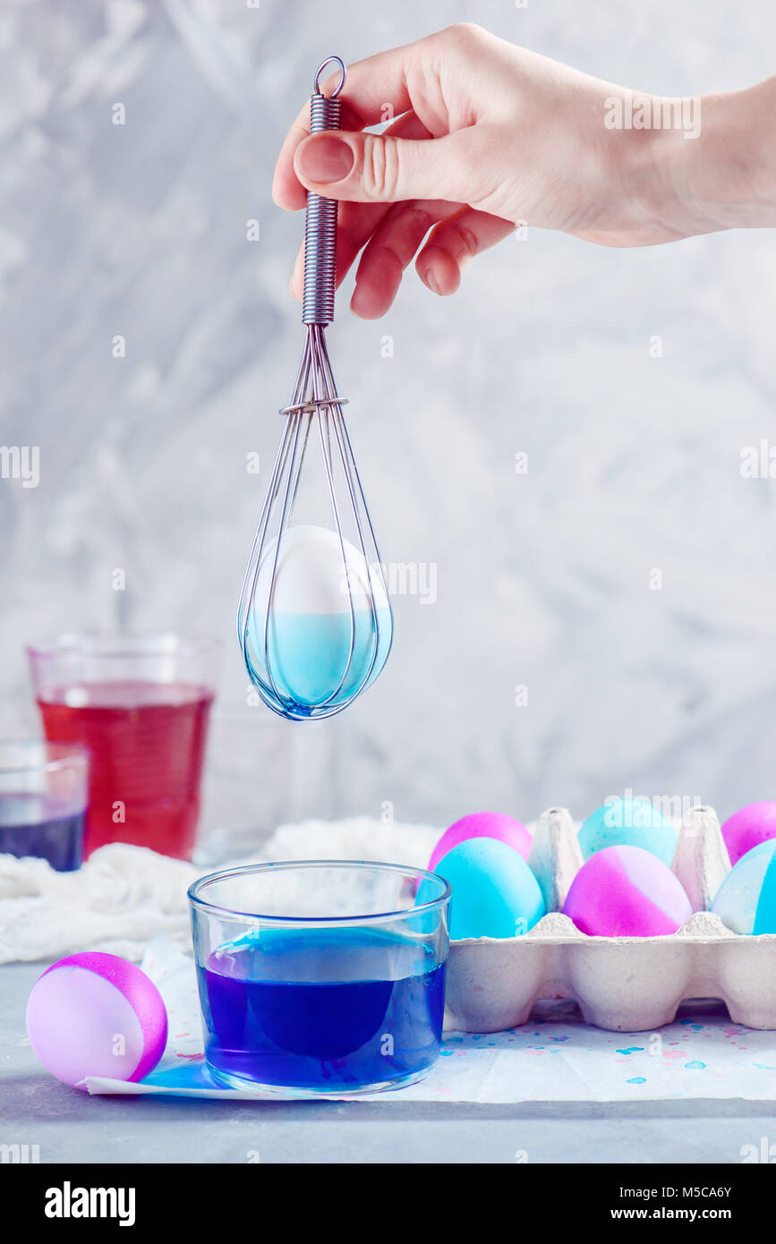 Easter egg in a whisk held by woman hand. Colorful holiday composition with food dye and paper egg tray. High key with copy space. Stock Photo