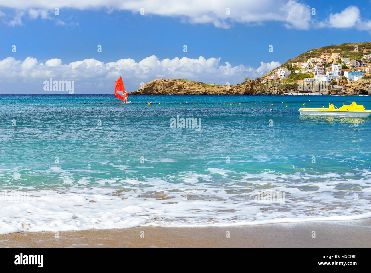 Windsurf with red sail rides on sea on background rocky shore. View from sunny Livadi beach in resort village Bali. Rethymno, Crete Greece. Extreme wa Stock Photo