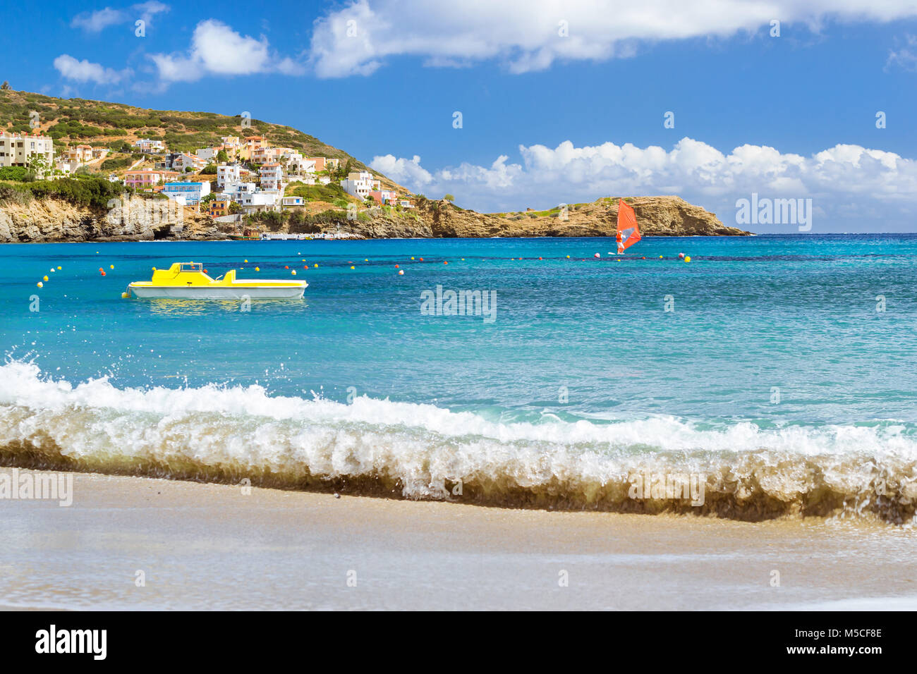 Windsurf with red sail rides on sea on background rocky shore. View from sunny Livadi beach in resort village Bali. Rethymno, Crete Greece. Extreme wa Stock Photo