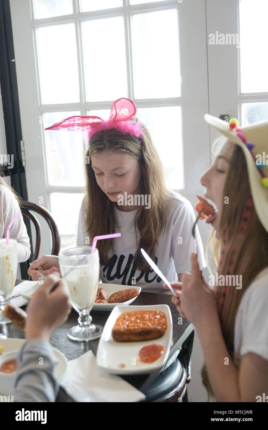 two young children are sitting at a party table eating fried food and drinking milkshakes- there are balloons and party decorations- they are talking Stock Photo