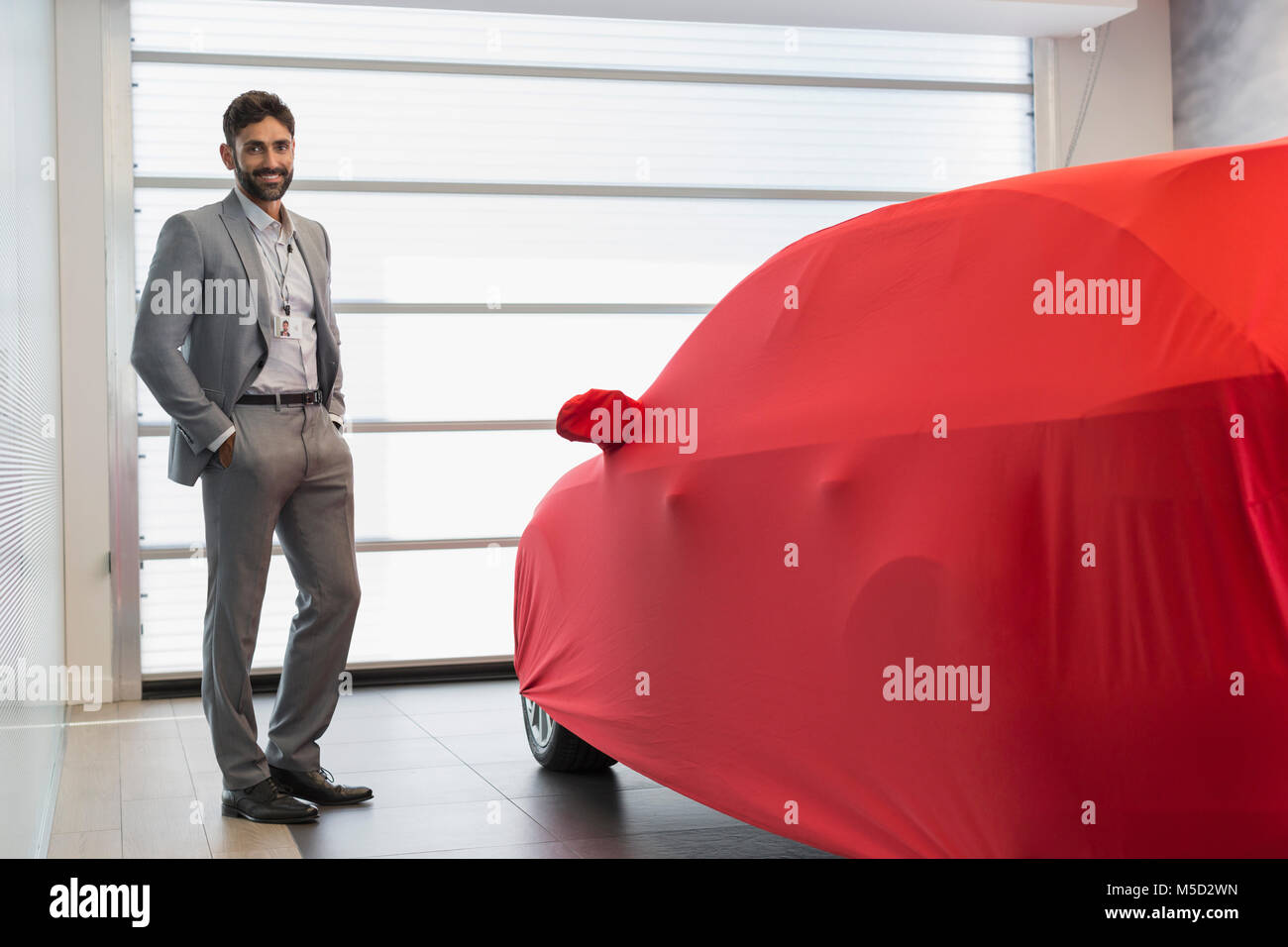 Portrait confident car salesman next to covered car in car dealership showroom Stock Photo