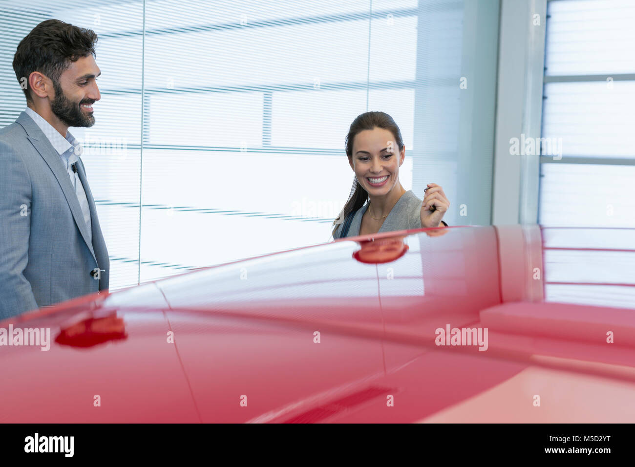 Smiling car salesman showing new red car to female customer in car dealership showroom Stock Photo