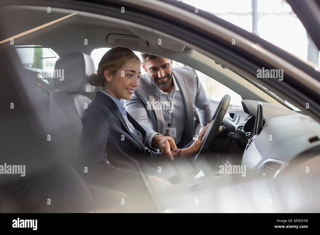 Car salesman explaining new car to female customer in driver’s seat in car dealership showroom Stock Photo