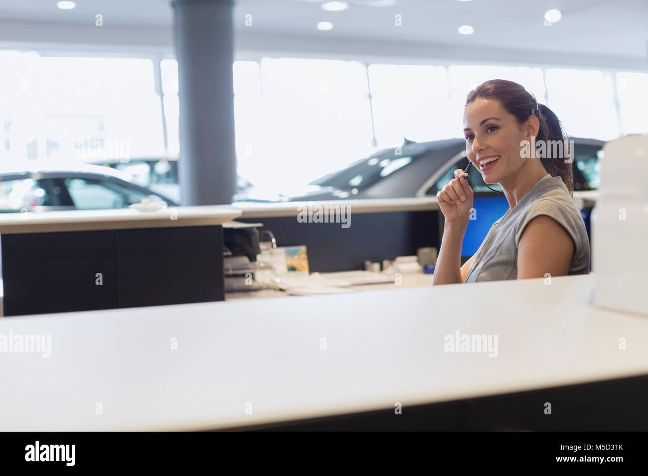 Smiling female receptionist talking on hands-free device telephone in car dealership showroom Stock Photo