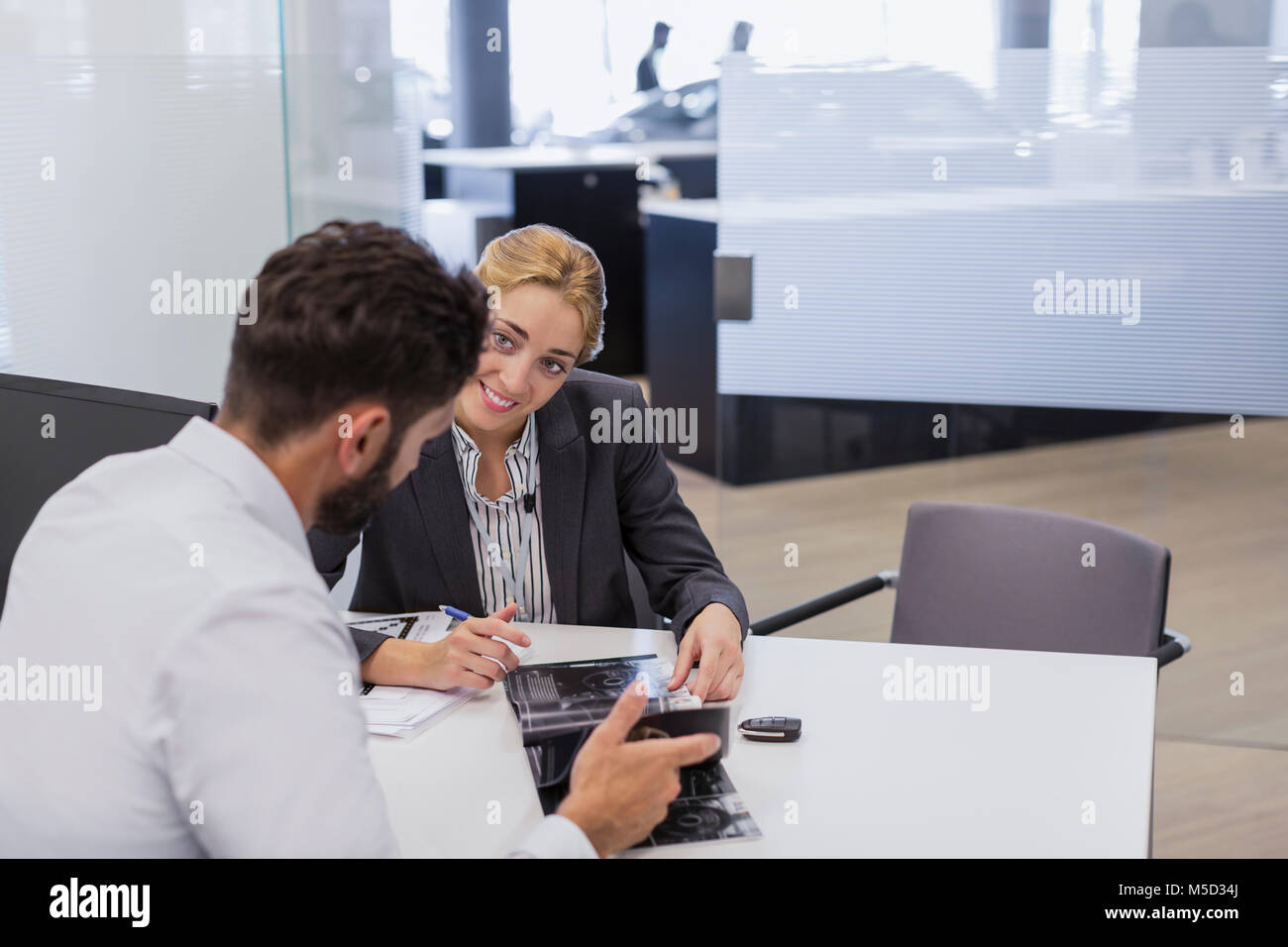 Smiling car saleswoman showing brochure to male customer in car dealership office Stock Photo