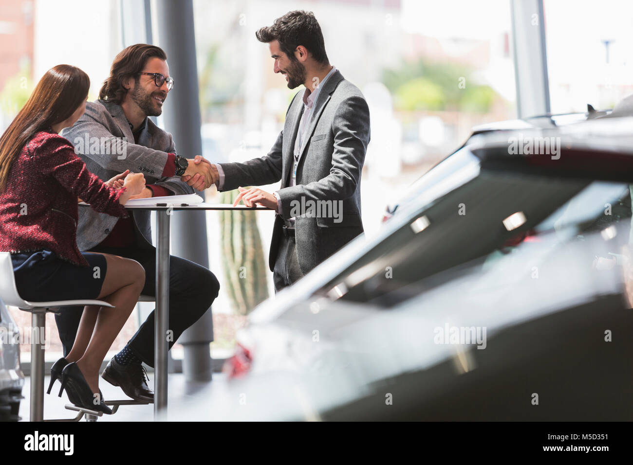Car salesman handshaking with couple customers at table in car dealership showroom Stock Photo