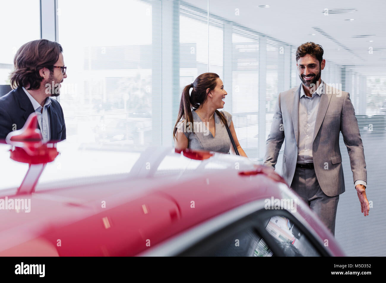 Car salesman and smiling couple customers looking at new car in car dealership showroom Stock Photo