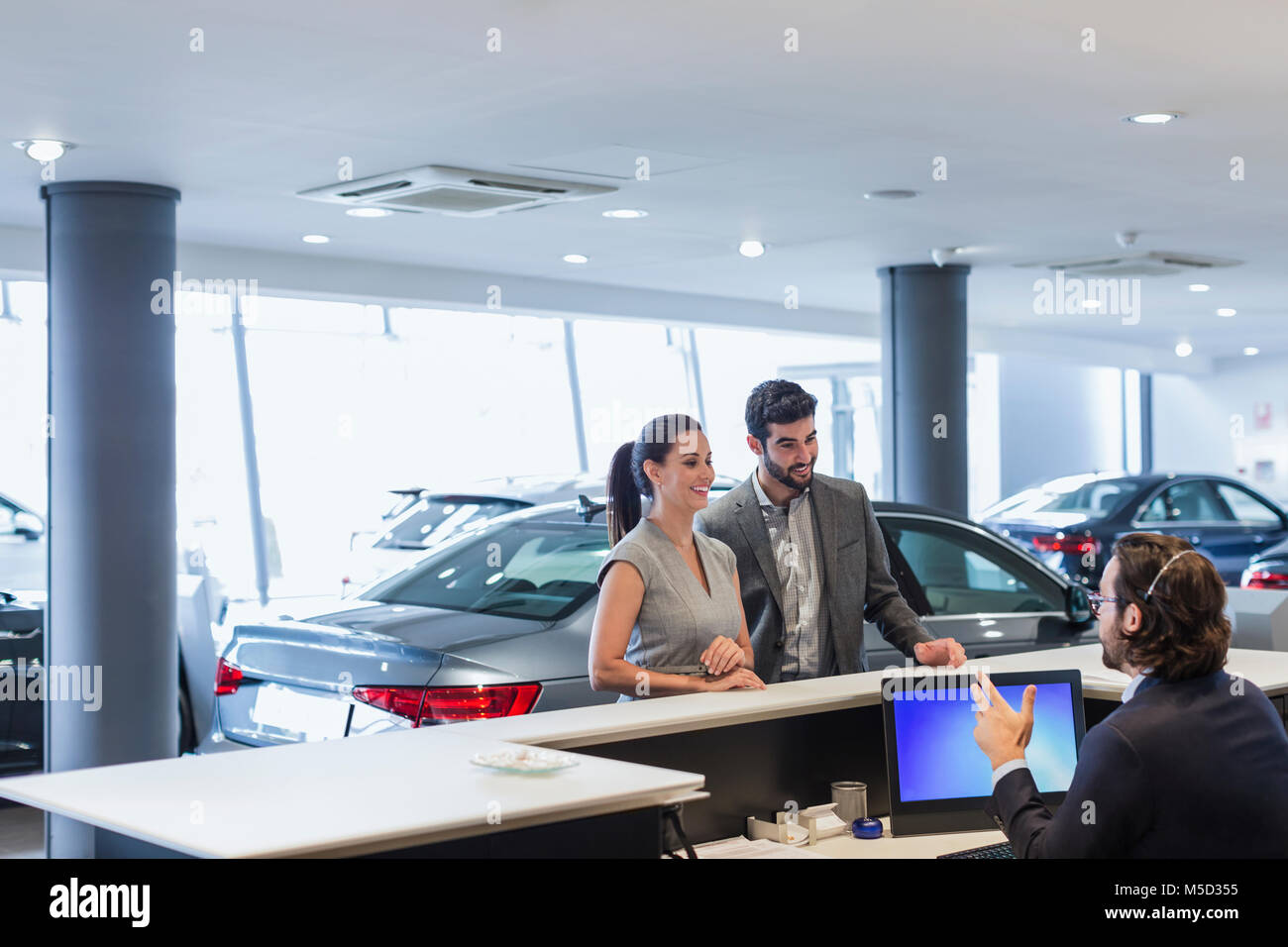 Couple customers talking to male receptionist at desk in car dealership showroom Stock Photo