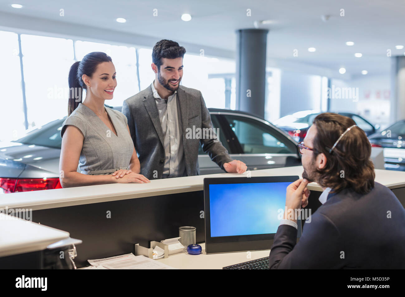 Couple customers talking to male receptionist at desk in car dealership showroom Stock Photo