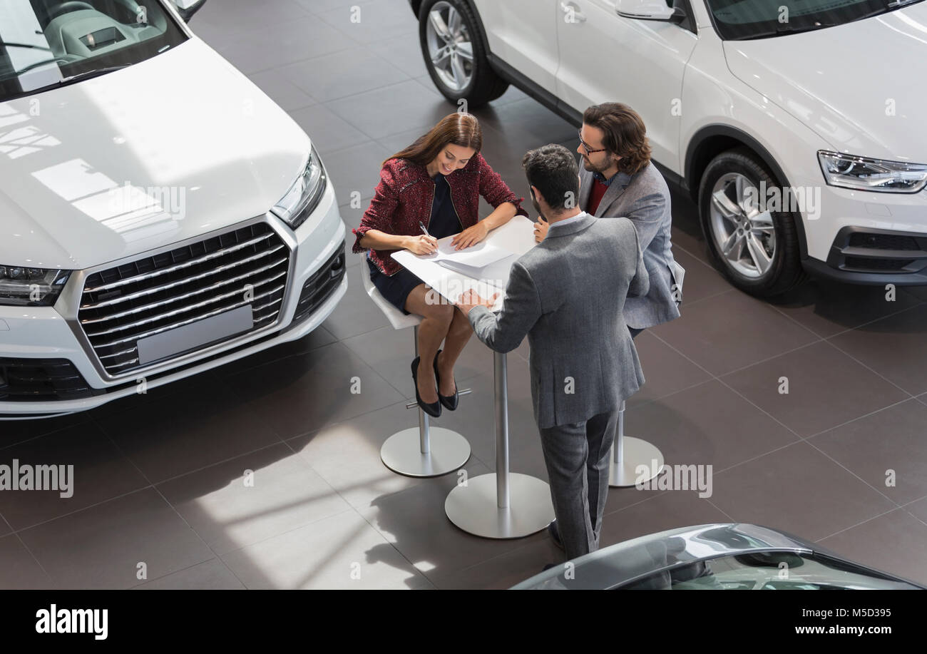 Car salesman watching couple customers signing financial contract paperwork in car dealership showroom Stock Photo