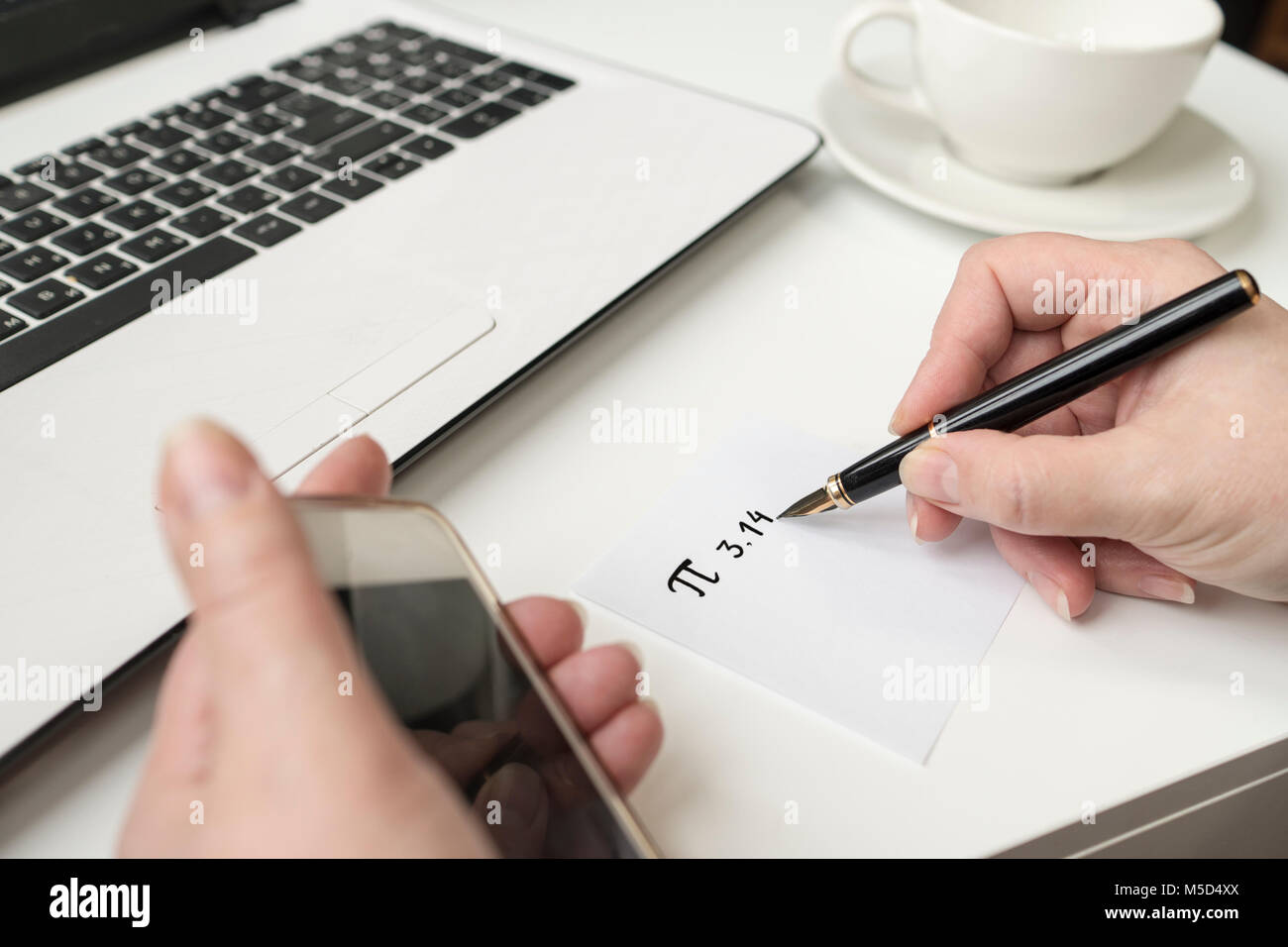 Happy Pi Day, written in black pen on white. On the desk a laptop, a cup, a hand holds the phone. Stock Photo