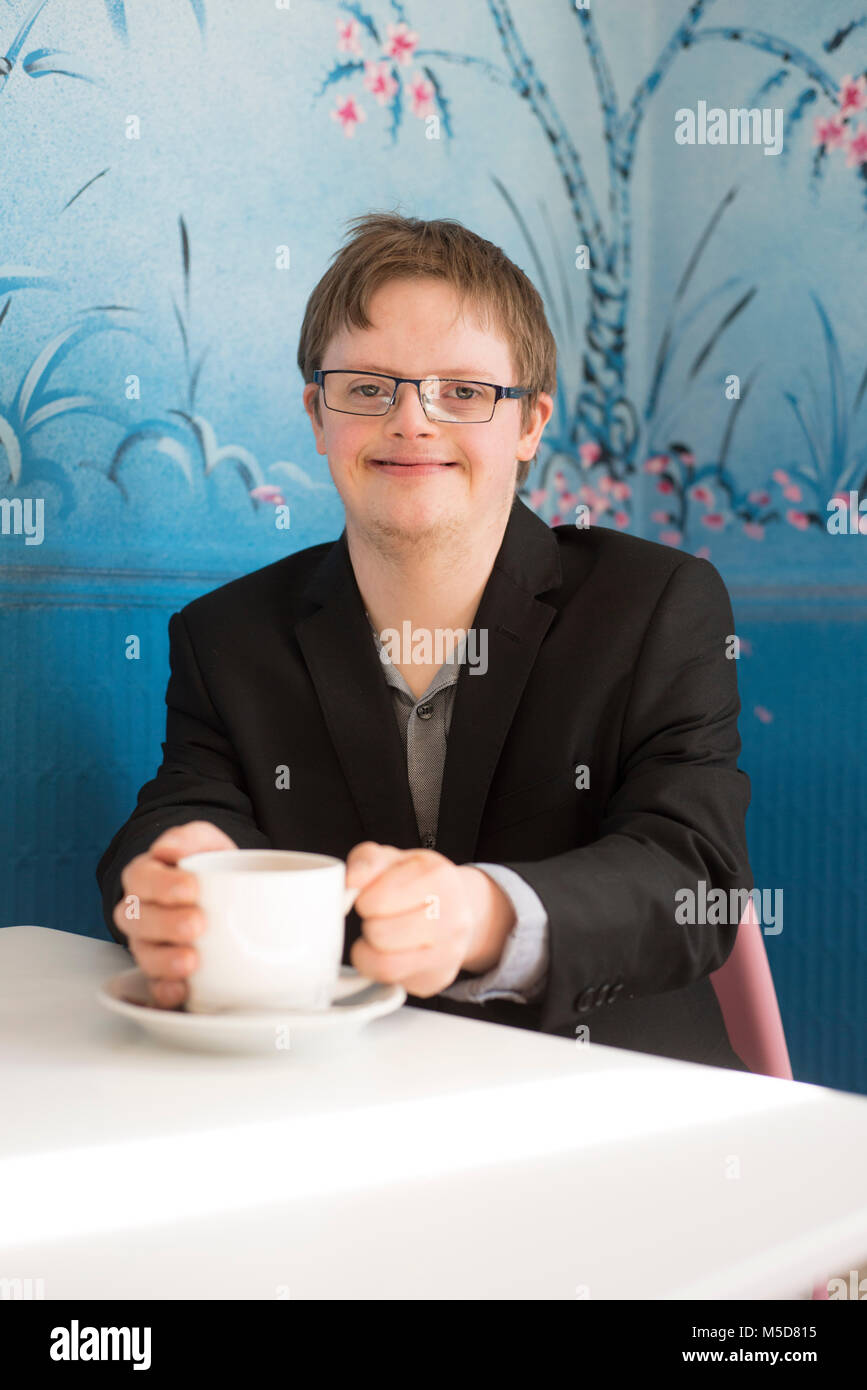 a young man with downs syndrome sits at a table in a cafe drinking coffee Stock Photo