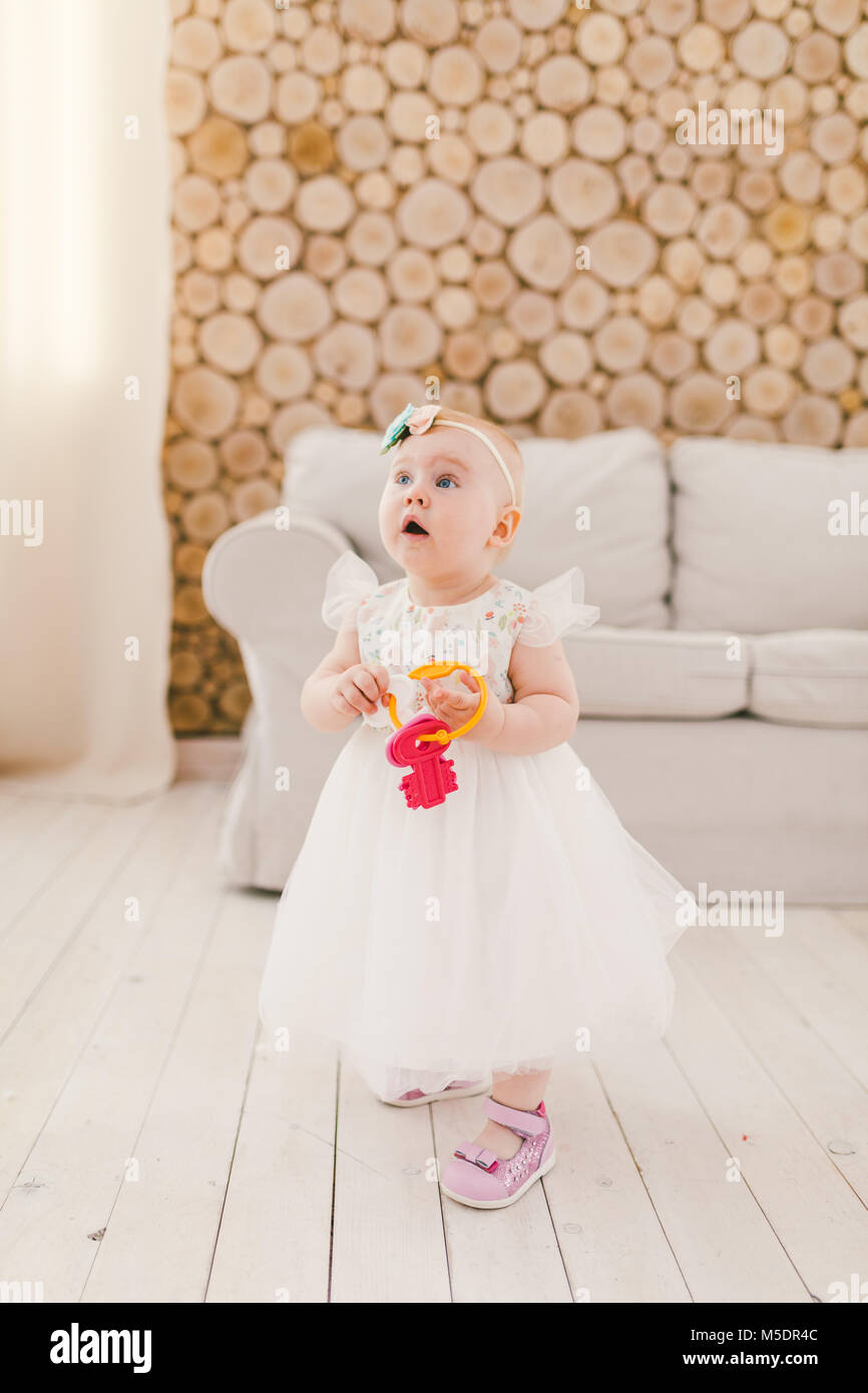 Little baby girl in white lavish dress standing in living room in house on sofa and wooden wall background and holding a bunch of toy plastic keys in  Stock Photo