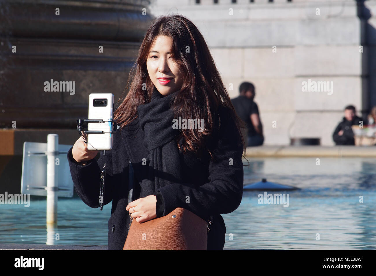 A young oriental woman taking a selfie in the winter sunshine next to a pool in Trafalgar Square, London using a selfie stick Stock Photo