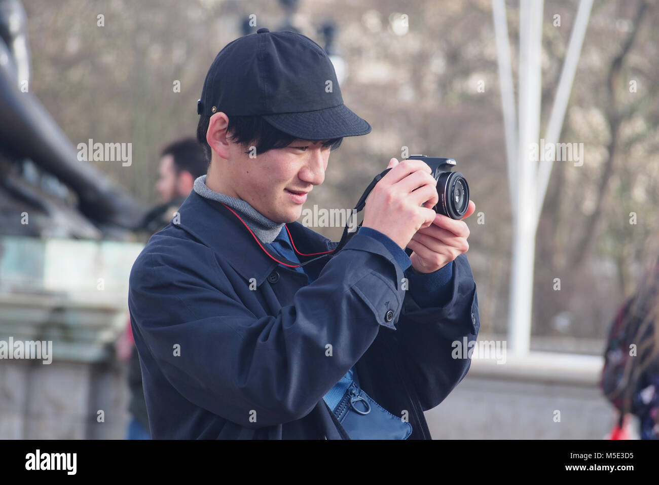 Japanese young man wearing a baseball hat and coat holding a camera composing a photograph on the Buckingham Palace Forecourt in the winter sunshine Stock Photo