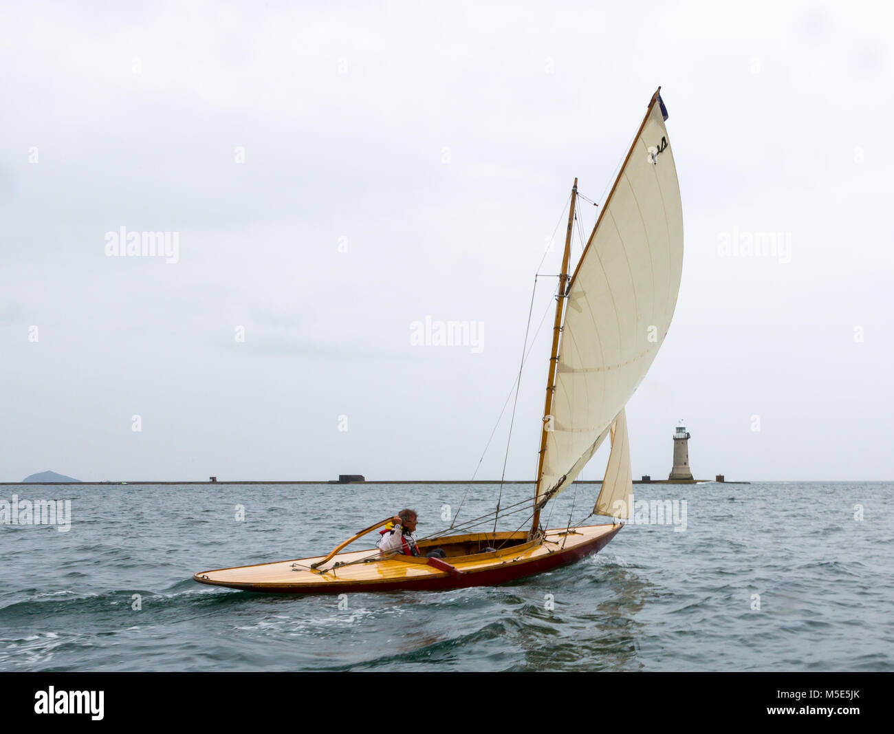 A small gaff rigged wooden yacht sails past Plymouth's Breakwater on an overcast day. Stock Photo