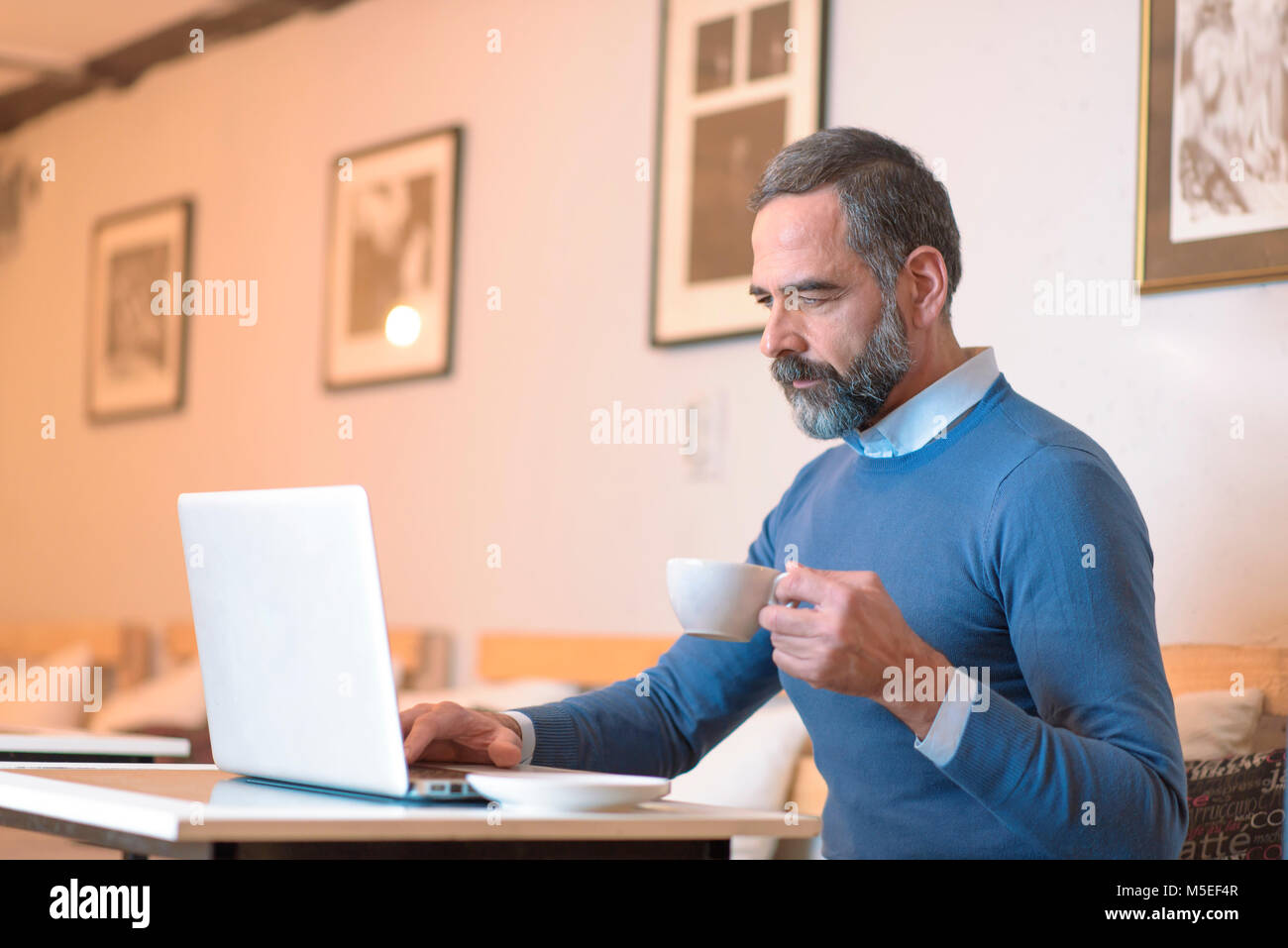 Casual senior man sitting in a coffee shop, having a coffee while browsing internet on his laptop Stock Photo