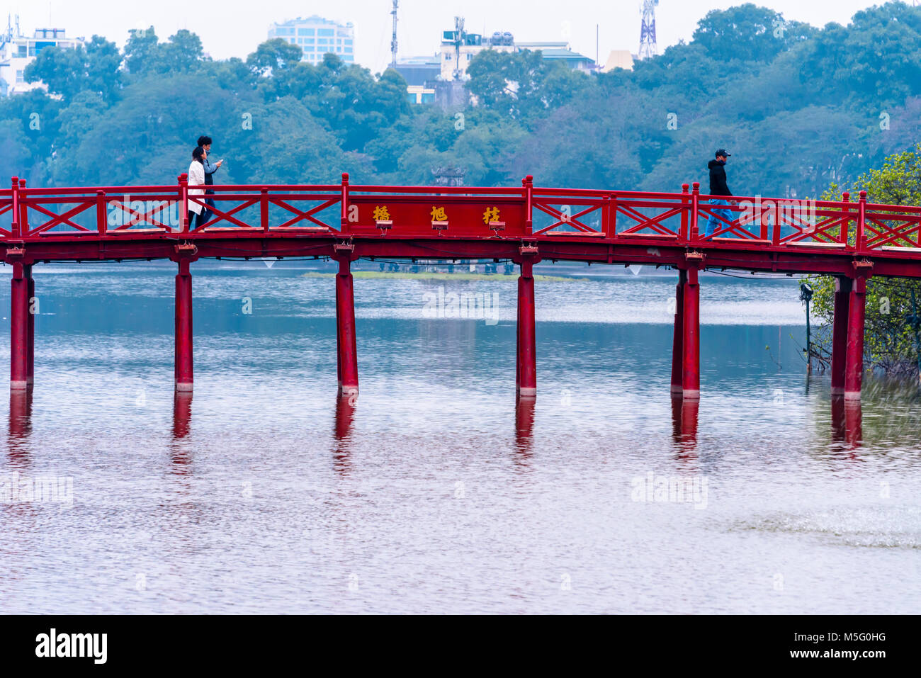 A young couple walk arcoss the iconic red painted Huc Bridge over Ho Hoan Kiem Lake, Hanoi, Vietnam which leads to the Den Ngoc Son Confucious Temple. Stock Photo