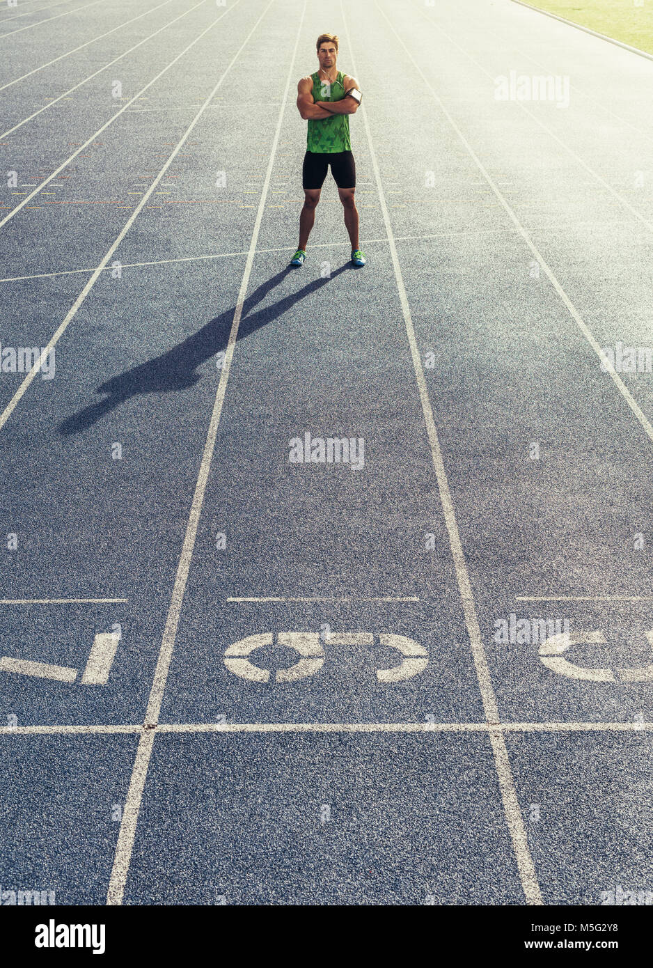 Runner standing on running track with hands folded. Athlete wearing earphones with mobile phone fixed in arm band. Stock Photo