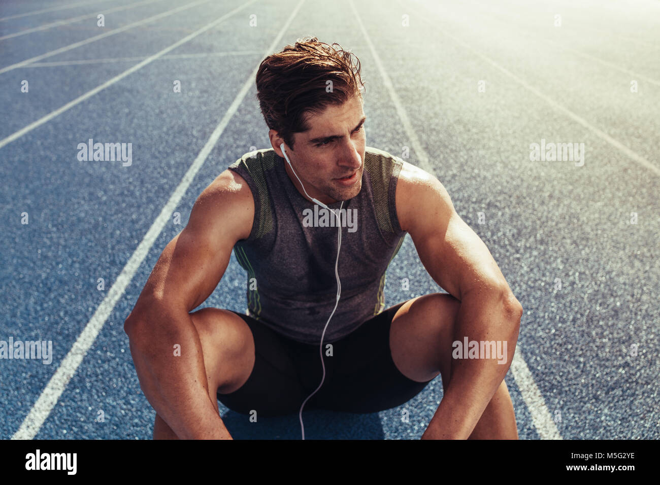 Close up of an athlete sitting on a running track listening to music. Runner wearing earphones sitting and relaxing on the running track. Stock Photo