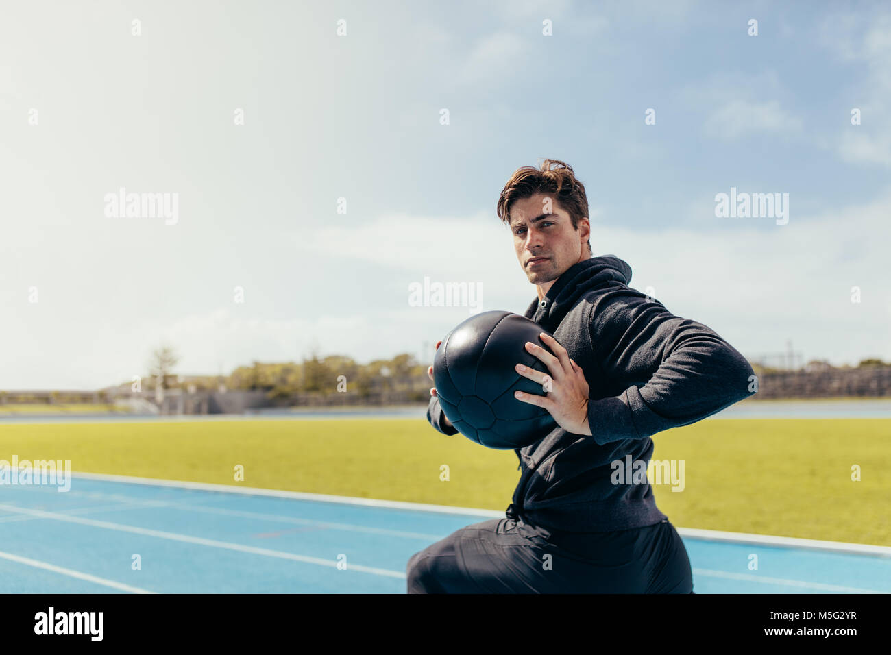 Runner doing a medicine ball workout on running track. Athlete using a medicine ball for fitness training at a track and field stadium. Stock Photo