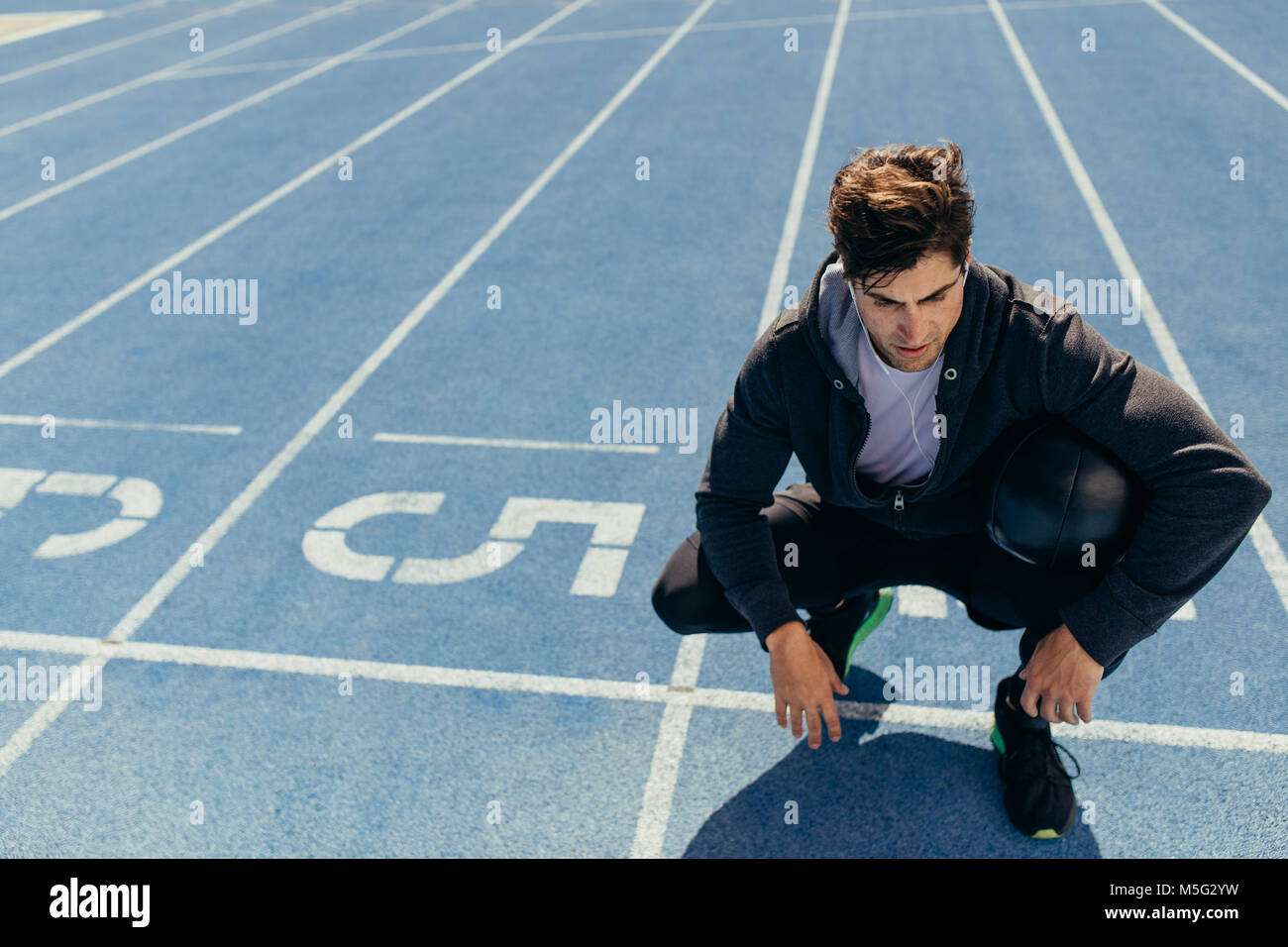 Athlete sitting on a running track near the start line with a medicine ball. Runner wearing earphones sitting on the running track holding a ball. Stock Photo