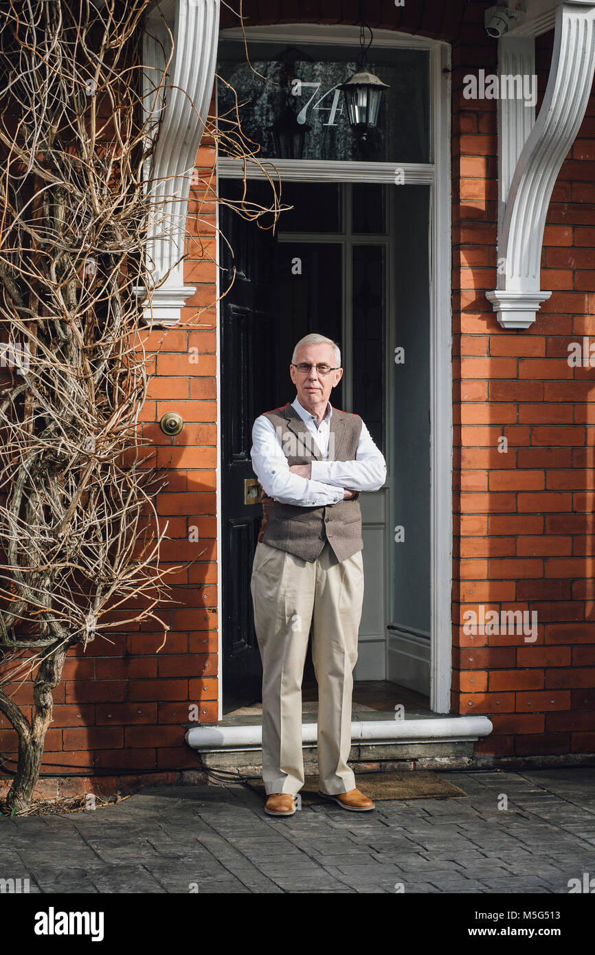 Happy senior man standing outside the front door of his home. He has his arms crossed and is looking at the camera. Stock Photo