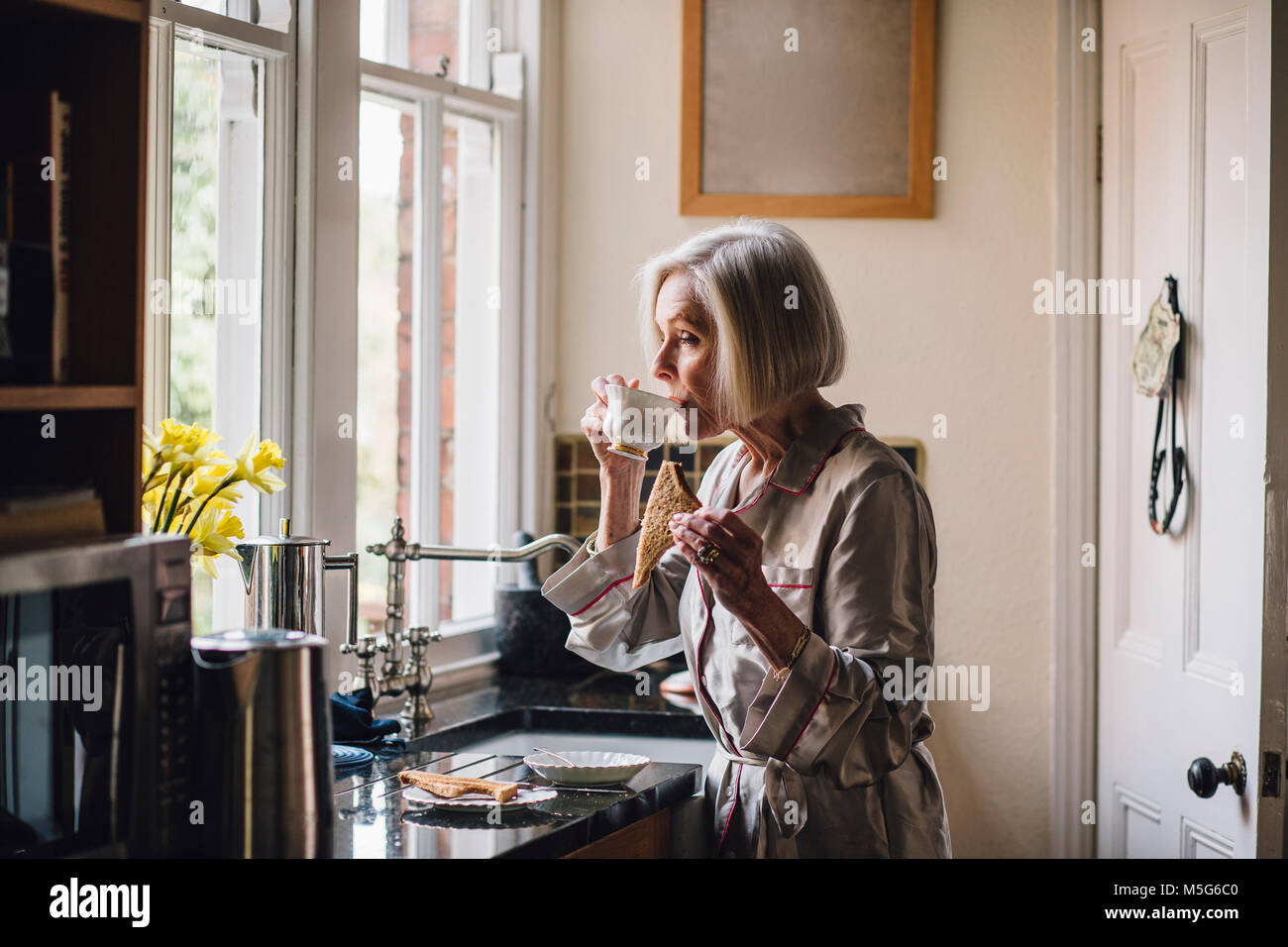 Senior woman is standing alone in her kitchen in her dressing gown, eating toast and dirnking tea. Stock Photo