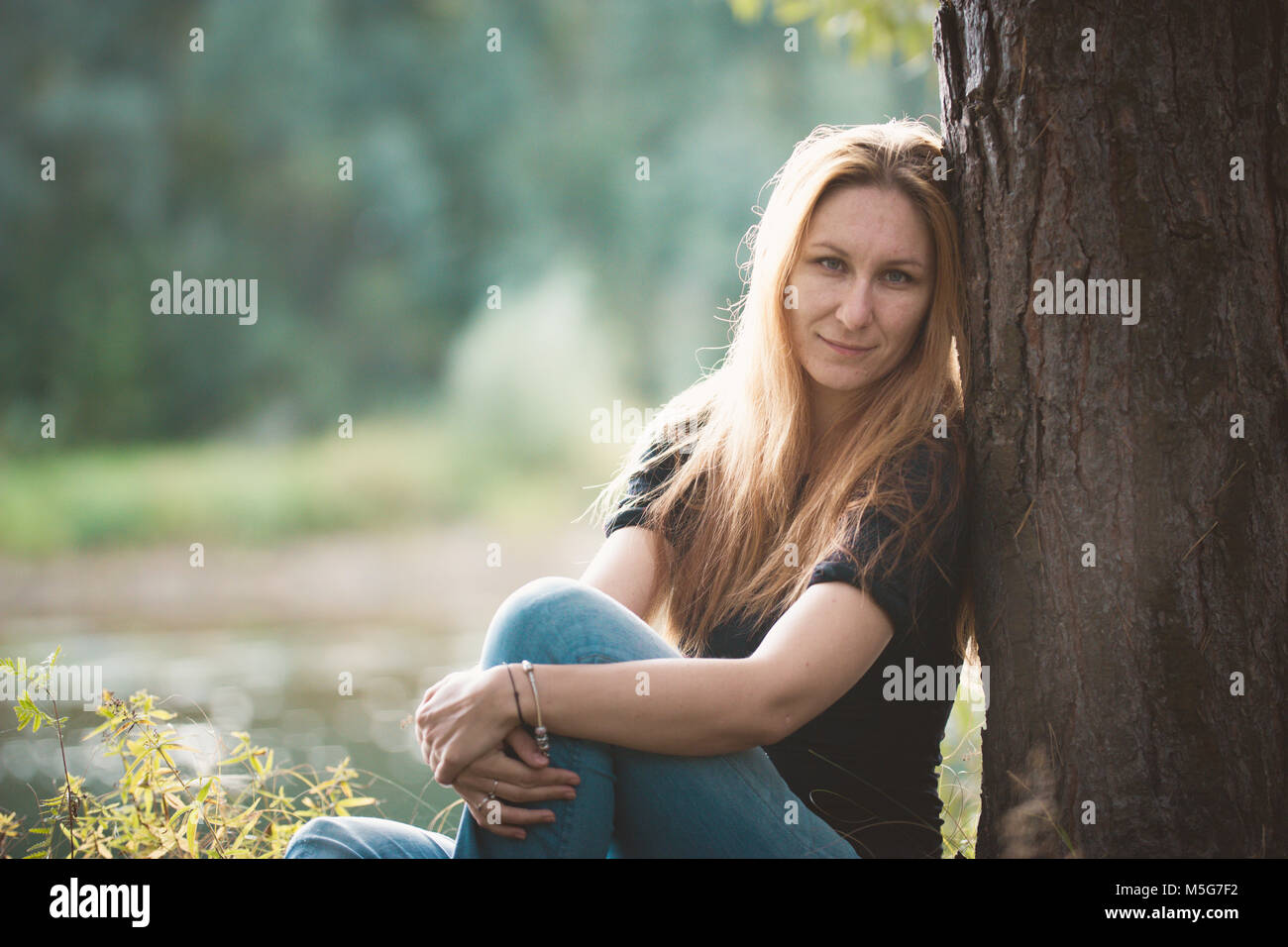 Beautiful red-haired girl sitting against a tree near river at forest ...