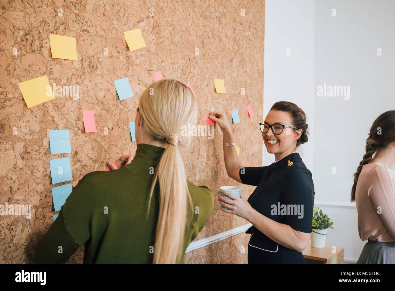 Two women are at work in an office. They are standing at a cork board and are having a discussion as they pin things up. Stock Photo