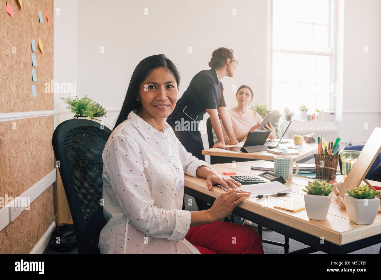 Portrait of a female employee in her workplace. She is sitting at her desk and is smiling at the camera. Stock Photo