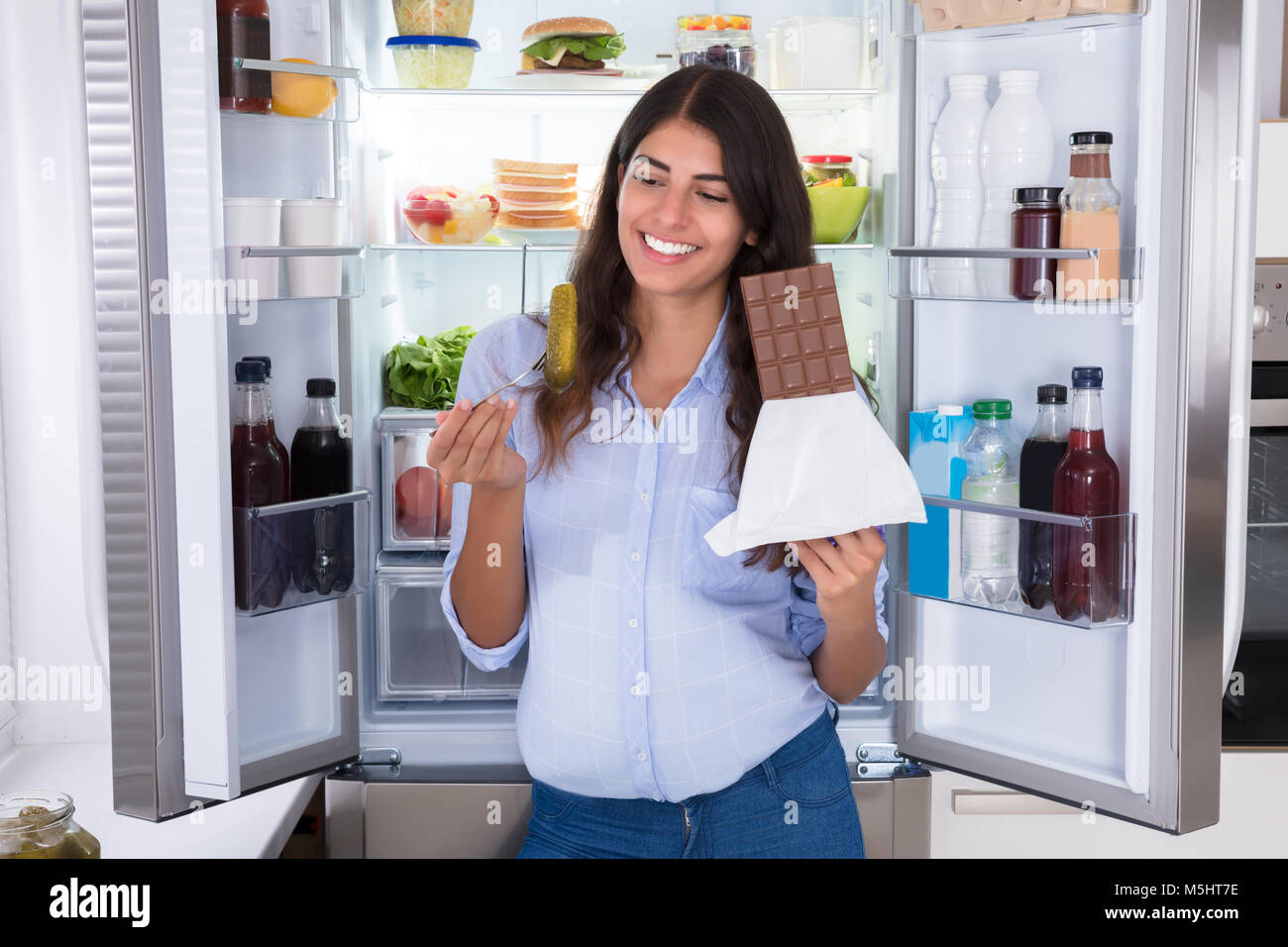 Young Woman Eating Chocolate Near Open Refrigerator Stock Photo