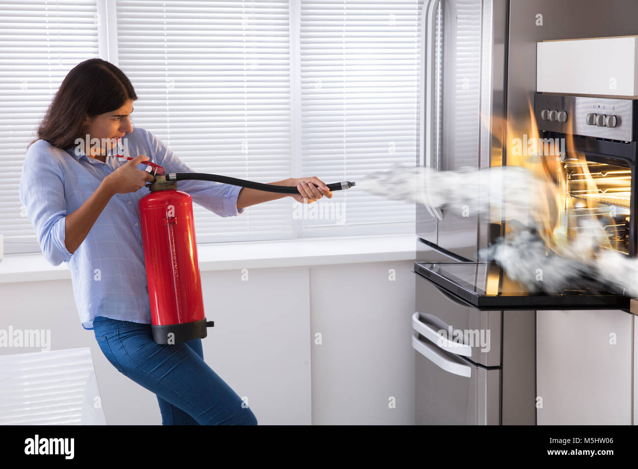 Young Woman Using Fire Extinguisher To Put Out Fire From Oven At Home