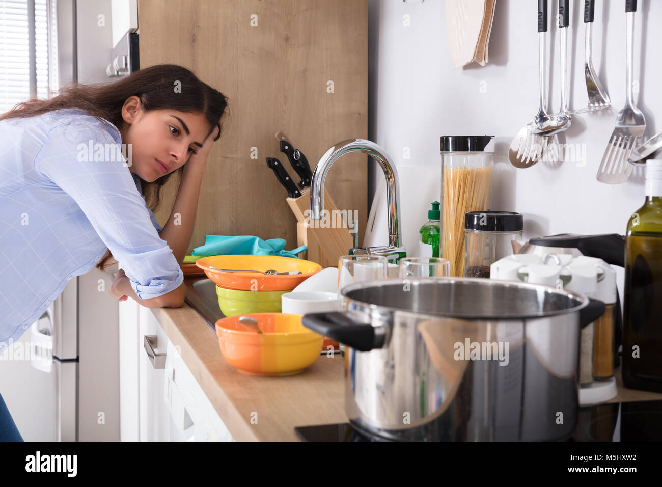 Tired Young Woman Standing Near Kitchen Sink Looking At Utensils In Kitchen Stock Photo