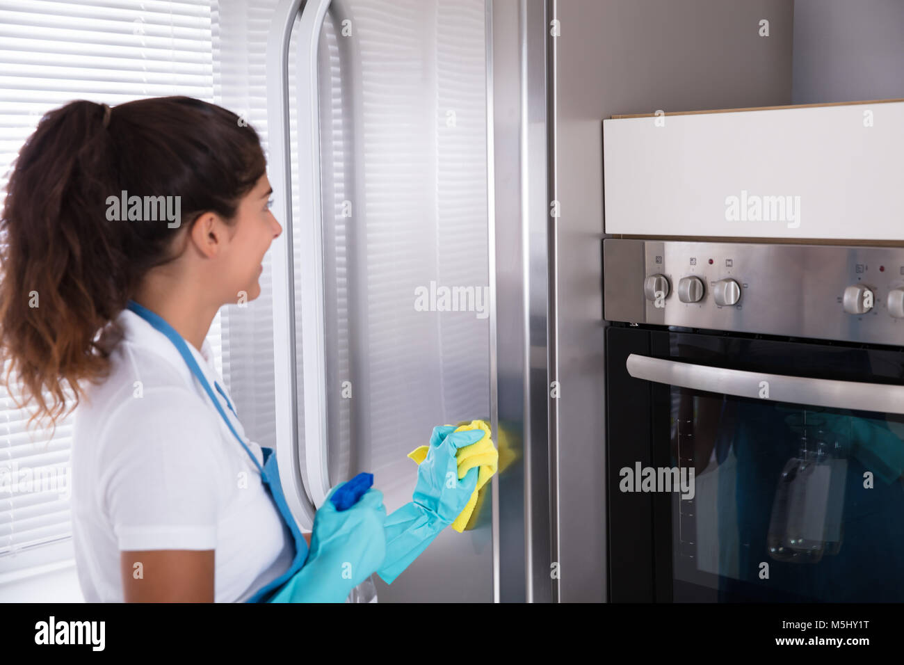 Happy Woman Cleaning Her Stainless Steel Refrigerator With A Cloth And Spray Bottle Stock Photo